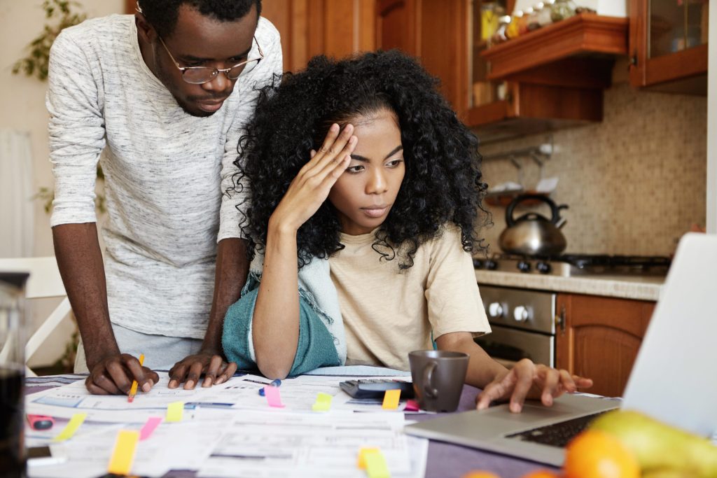 Serious young dark-skinned female with Afro hairstyle sitting in front of open laptop, using online banking app to review family accounts while analyzing domestic budget together with her husband